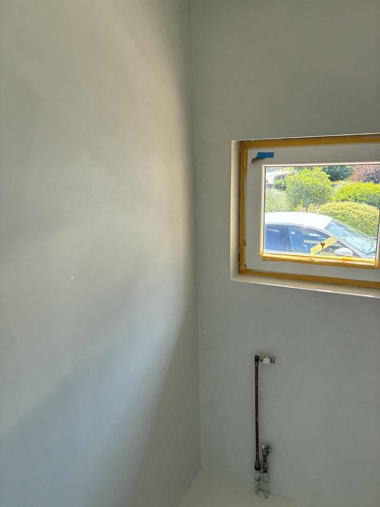 Innenraum eines frisch verputzten Raumes mit einem Fenster und sichtbarer Wasserleitung.