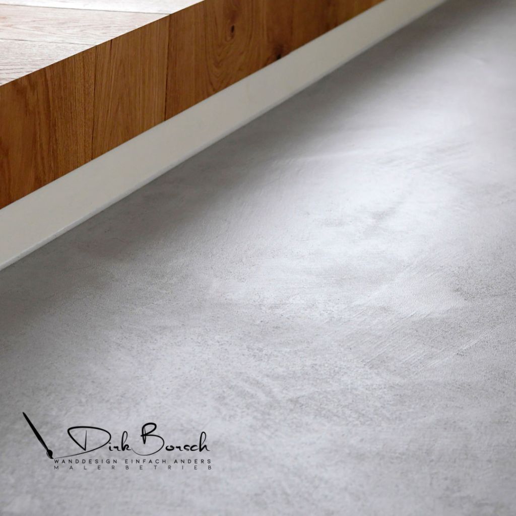 Detailaufnahme eines Fußbodens mit grauem Betonfinish und einer Holzoberfläche im Hintergrund.