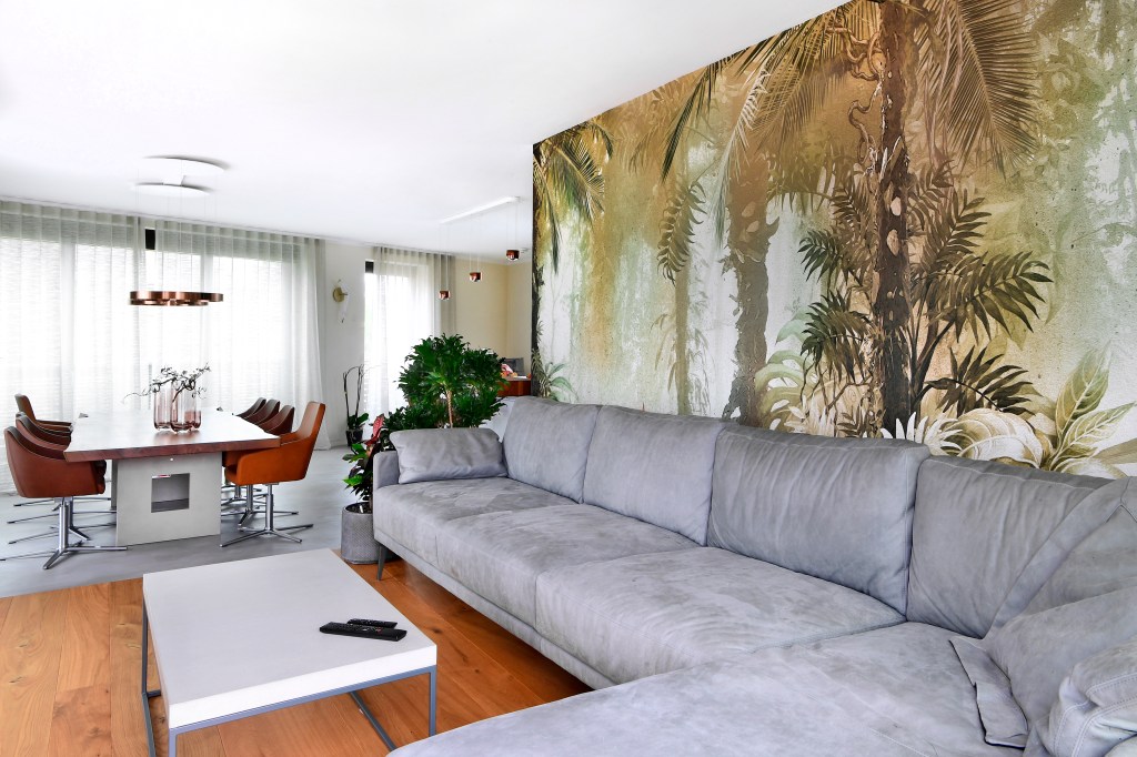 Ein modernes Wohnzimmer mit einem grauen Sofa, einem Couchtisch und einer Dschungel-Wandtapete. Im Hintergrund befindet sich ein Esstisch mit Stühlen und großen Fenstern, die viel Tageslicht hereinlassen.