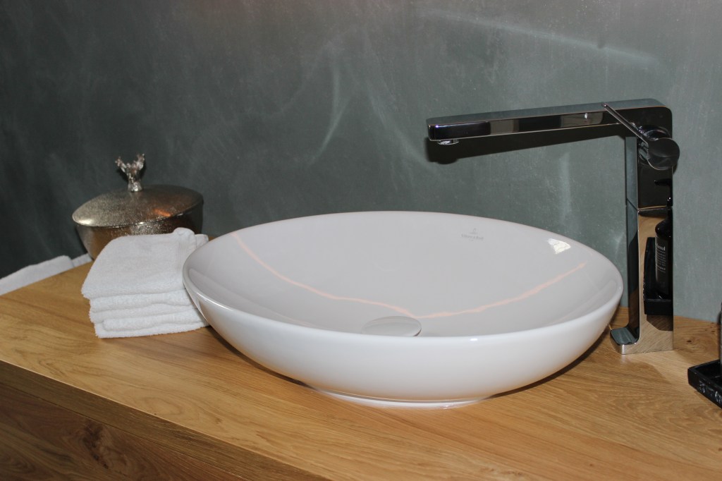 Ein modernes Waschbecken aus weißem Keramik auf einer Holzfläche, neben einem Stapel Handtücher und einer dekorativen Box.