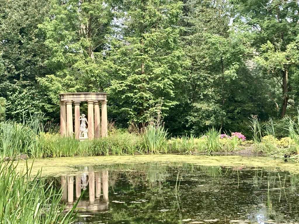 Ein ruhiger Garten mit einem Teich und einer Statue in einem Säulengebäude, umgeben von üppigem Grün und Blumen.